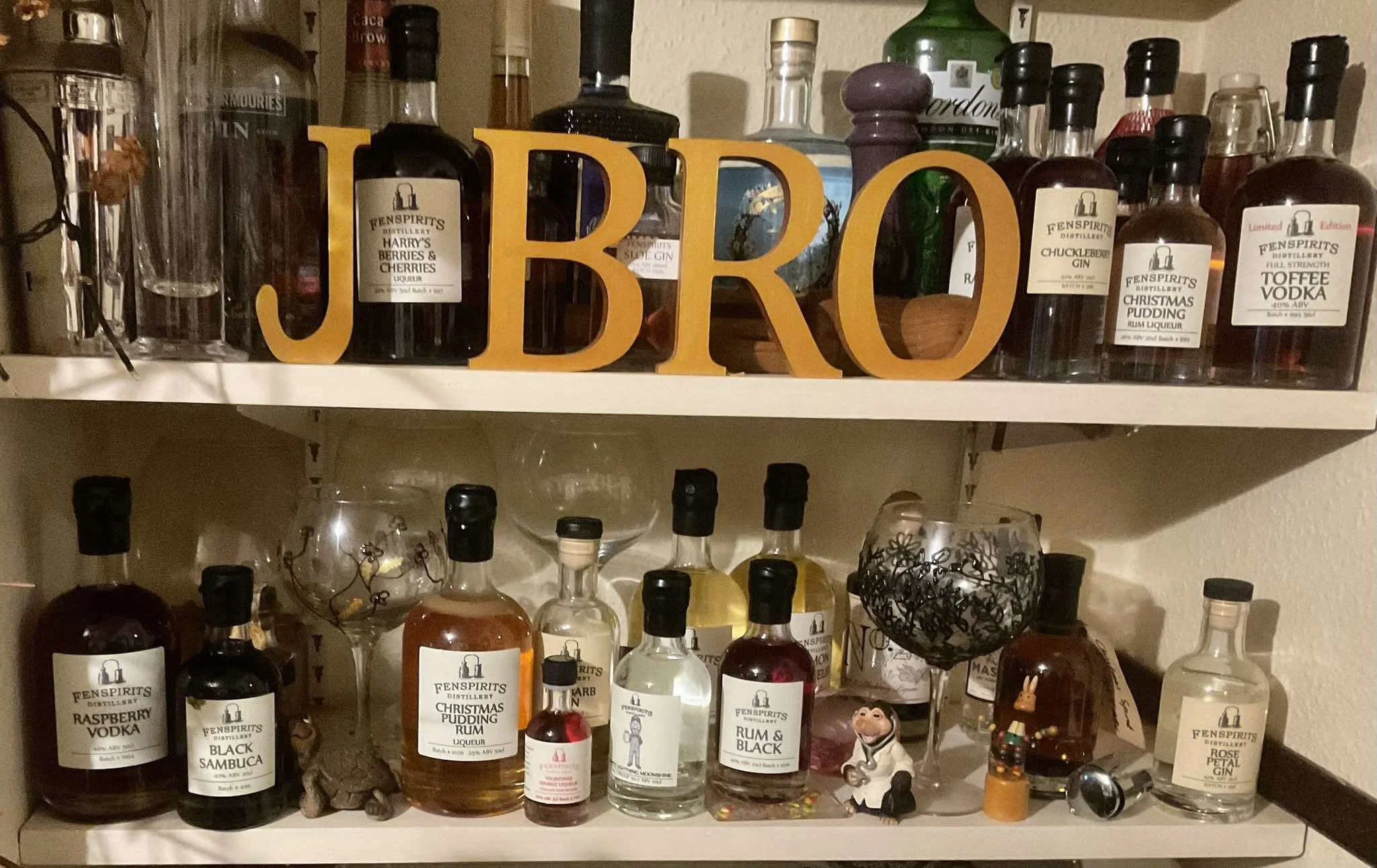 Shelf with various bottles of alcohol including vodka, rum, gin, and liqueurs, with yellow wooden letters spelling 'UBRO' on top. There are decorative items, including figurines, interspersed among the bottles.