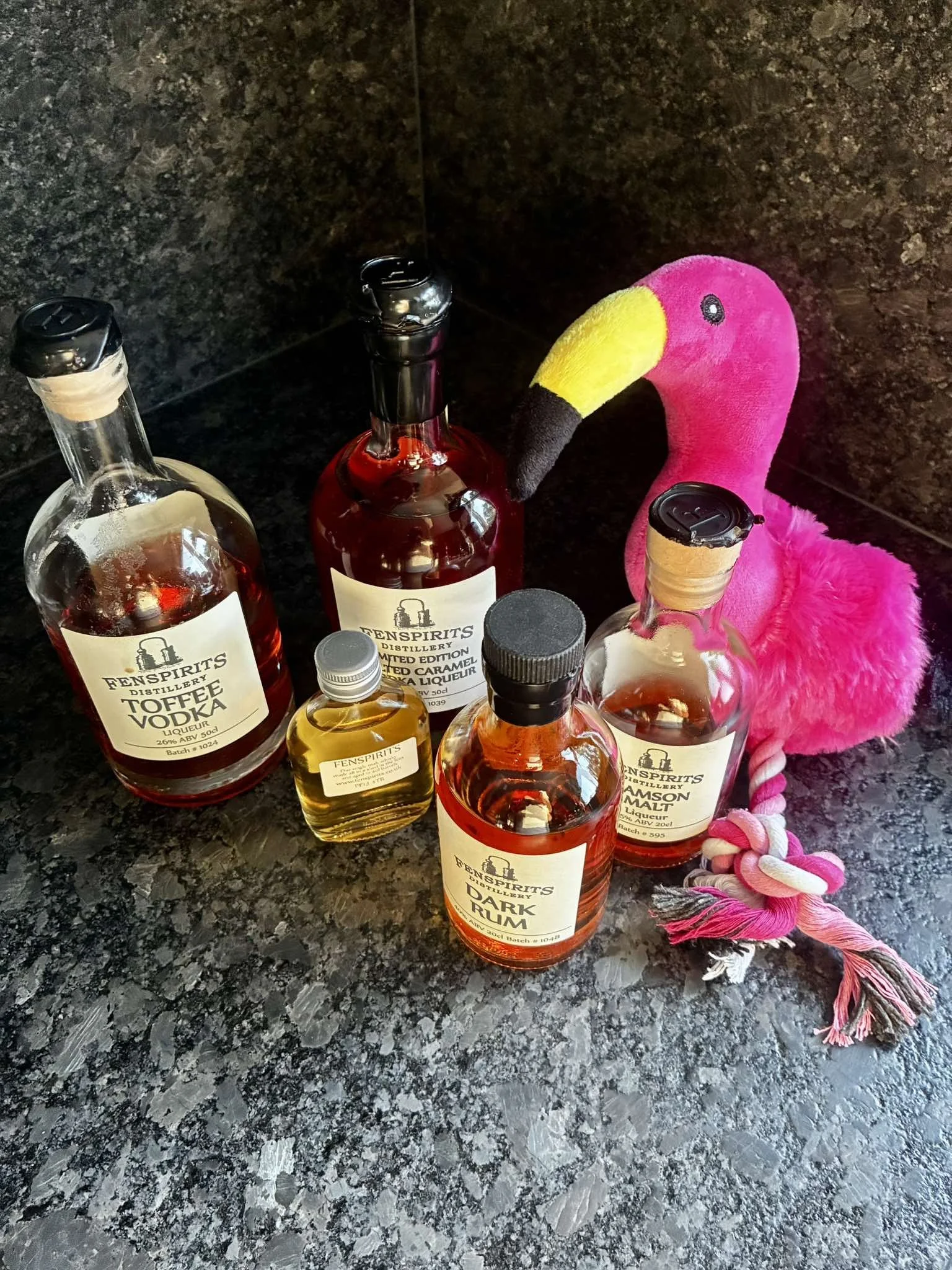 Various bottles of flavored liquor on a black granite countertop, with a pink plush flamingo drinking from one of the bottles.