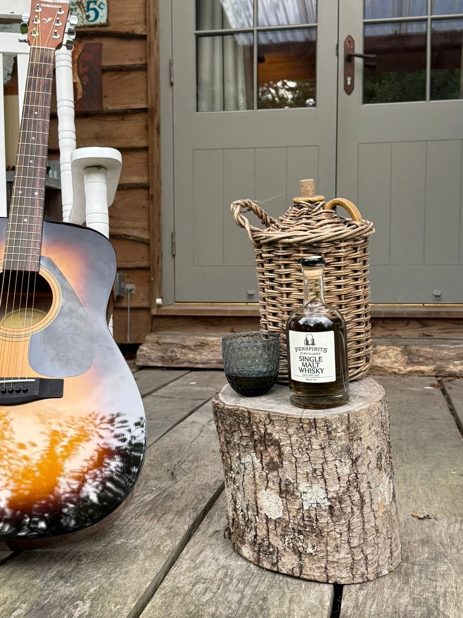 A guitar leaning against a white chair on a wooden deck, with a wooden wall and a glass door in the background. On a tree stump, there is a bottle of Fenspirits single malt whisky, a black glass, and a wicker basket.
