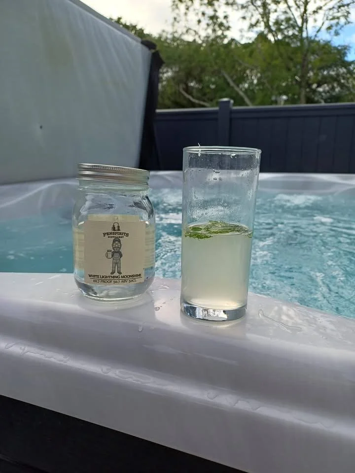 A jar labeled 'White Lightning Moonshine' and a tall glass of a lime-infused drink on a hot tub ledge outdoors.