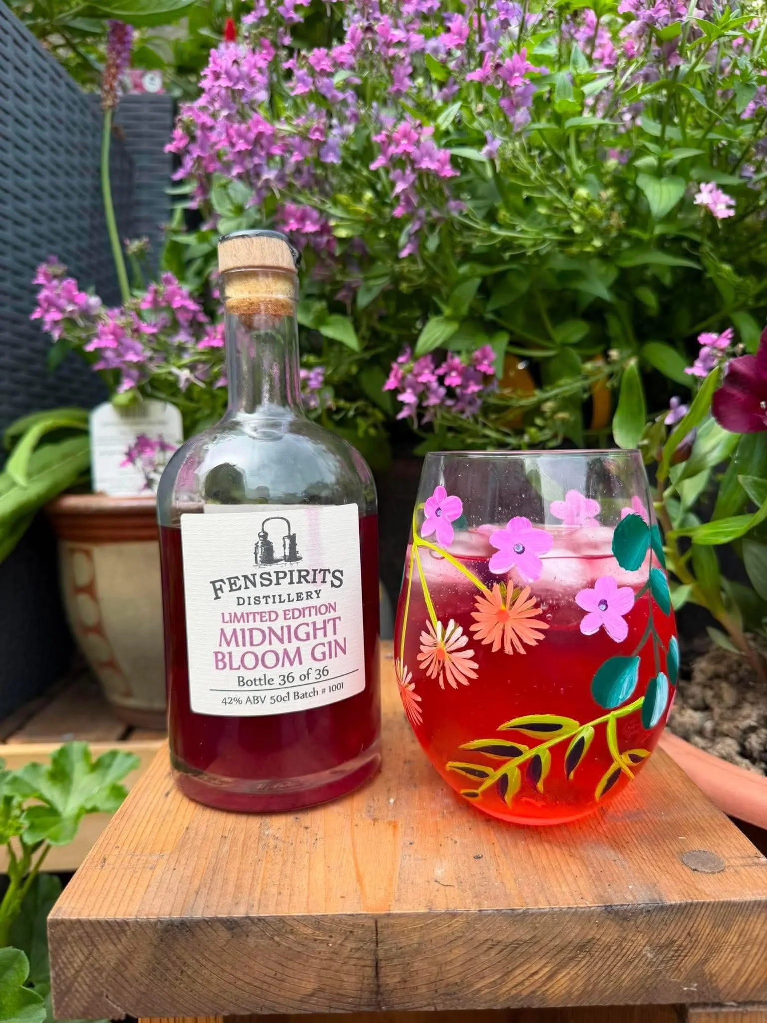 A bottle of Fenspirit Midnight Bloom Gin and a colorful flower-patterned drink glass on a wooden surface, with pink and purple flowers and green foliage in the background.