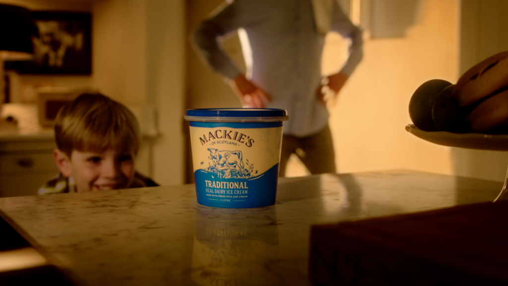 A boy smiling in a kitchen with a tub of Mackie's traditional real dairy ice cream on the counter in front of him, and a man with his hands on his hips in the background.