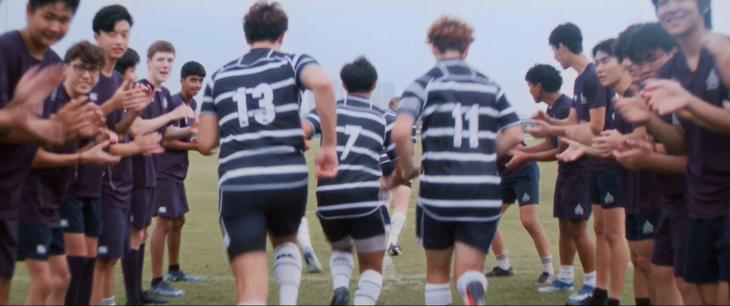 A group of young soccer players in striped uniforms running onto the field through a tunnel of teammates clapping on either side during daylight.