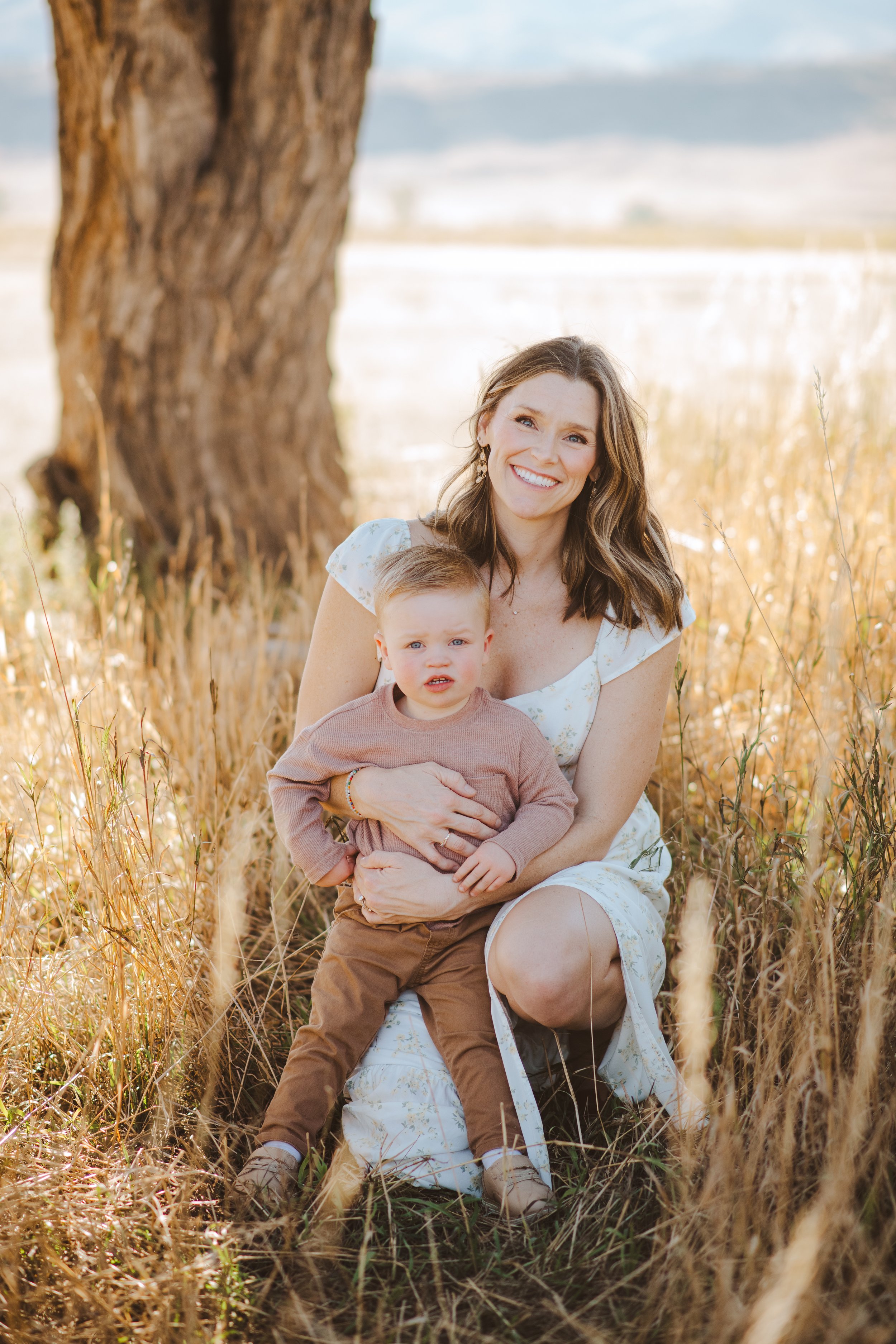 woman with brown hair, smiling and holding her toddler in a field.