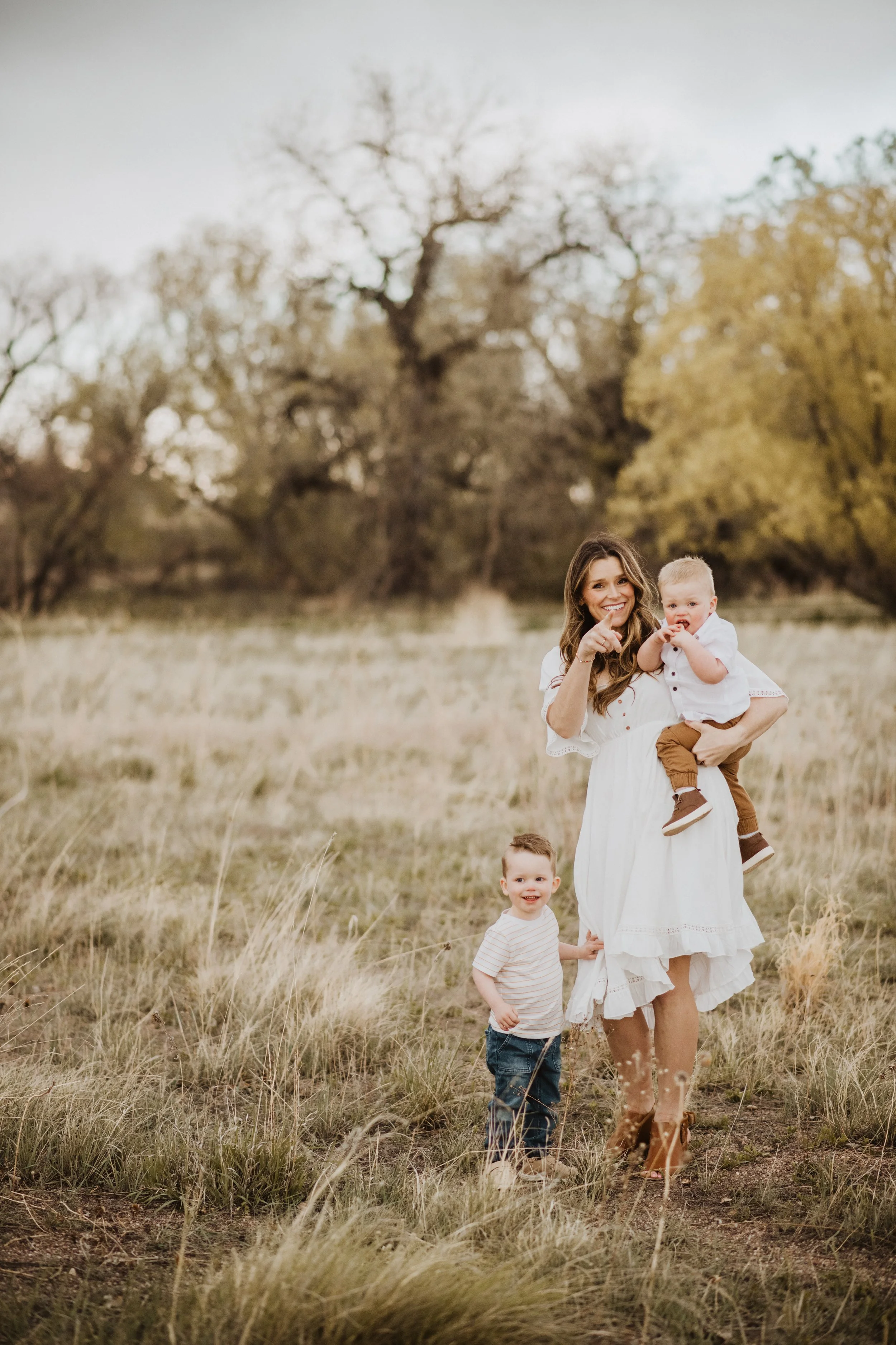 woman with long brown hair in white dress, holding a littl blonde toddler with another toddler holding onto her dress, in a field, smiling and pointing at the camera