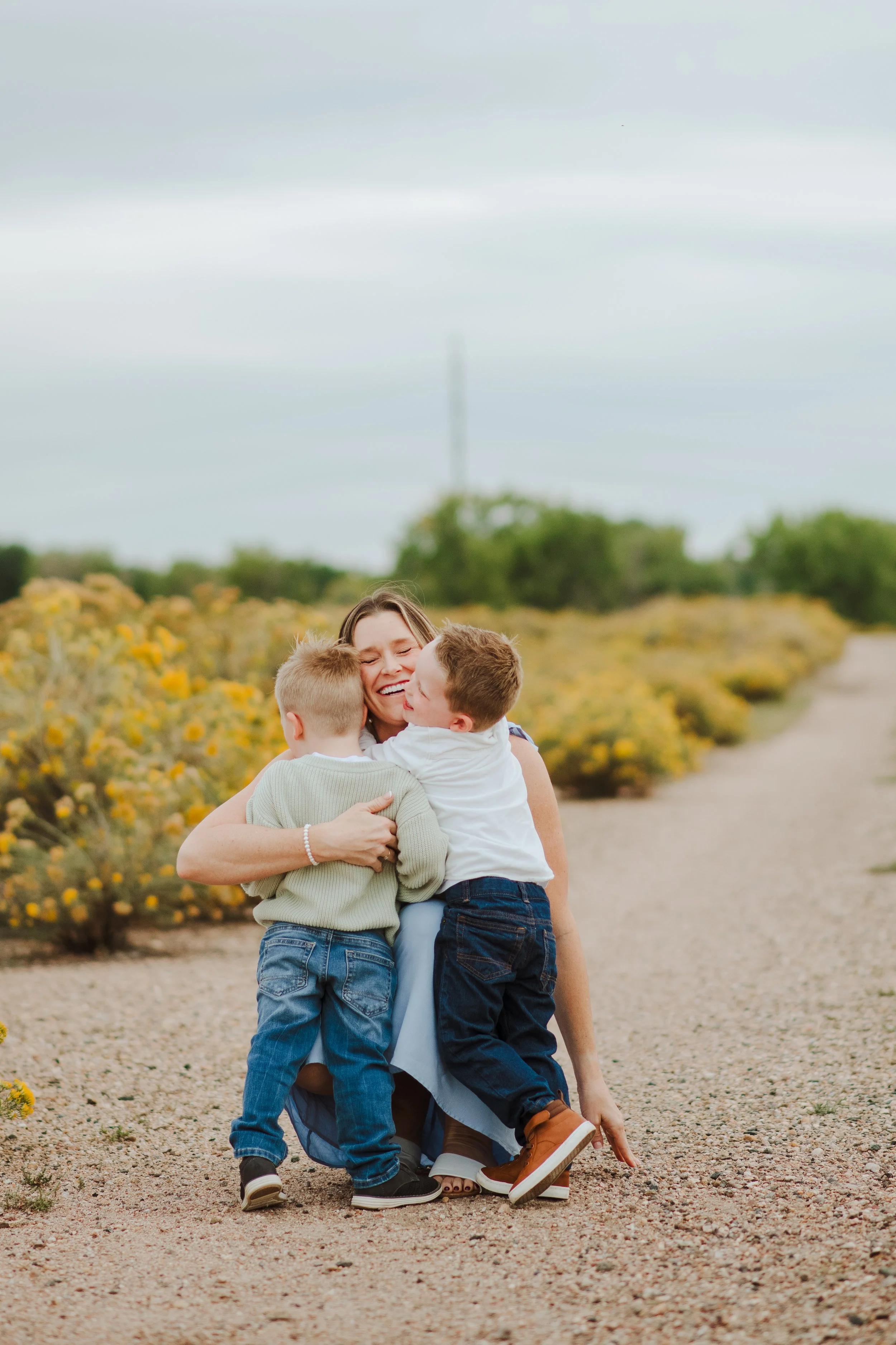 women with light brown hair in a blue dress smiling with her eyes shut while she hugs her two young boys, wearing a green sweater and a white button up with jeans and brown shoes on a gravel road