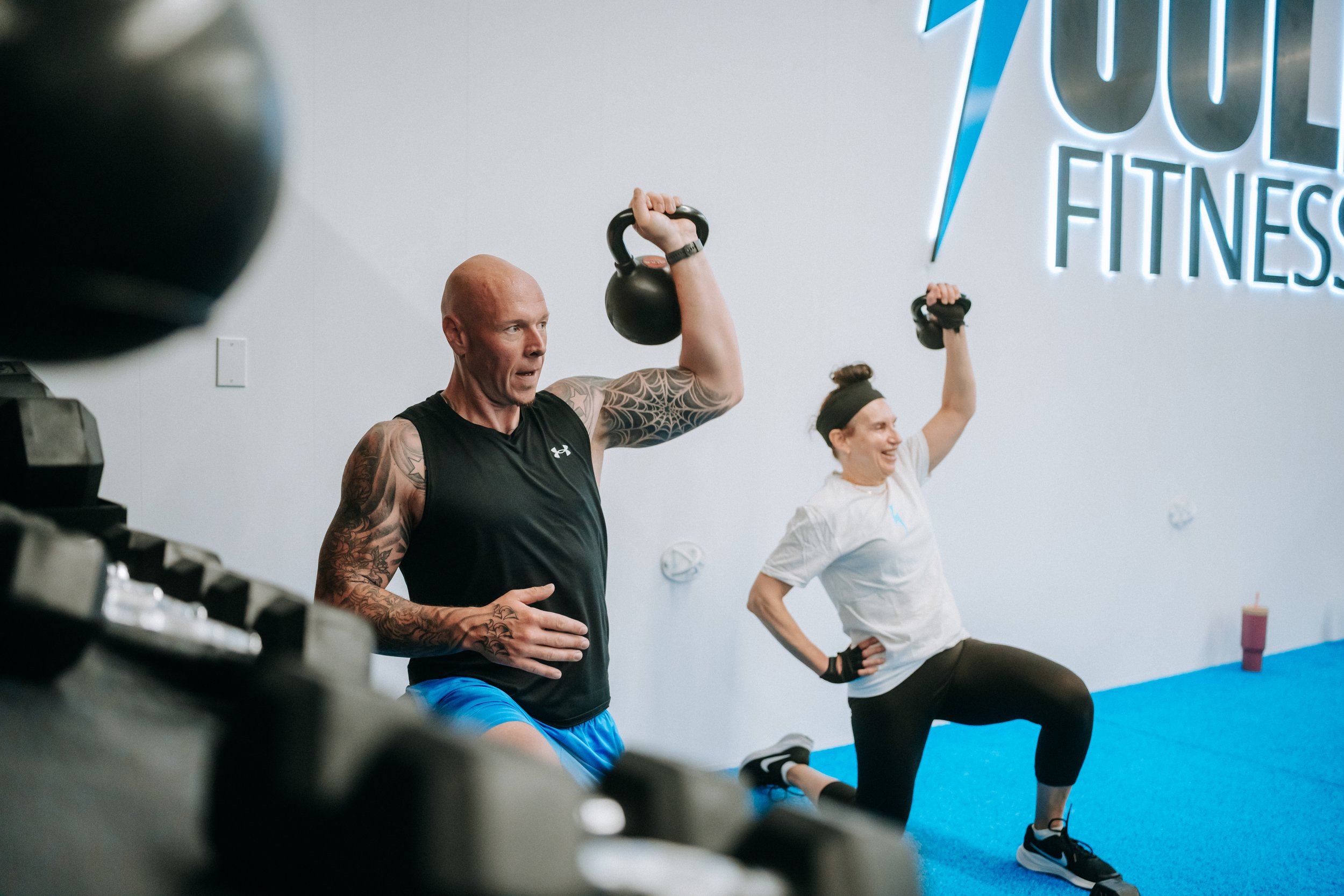 Coach Charlie leading a half-kneeling kettlebell press beside a smiling member at Jolt Fitness near Exeter, Ontario
