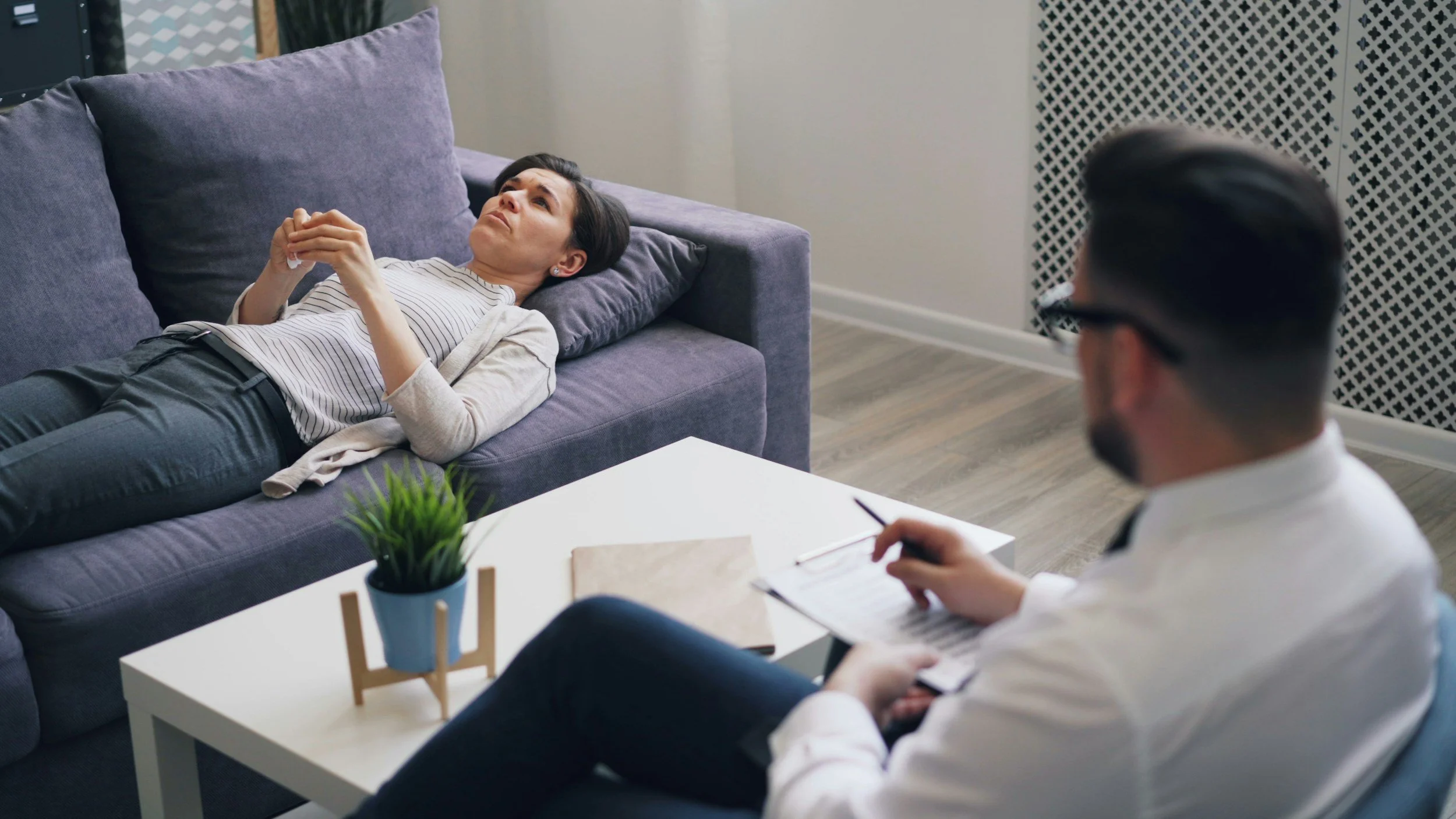 A woman lying on a purple sofa during a therapy session with a therapist taking notes.