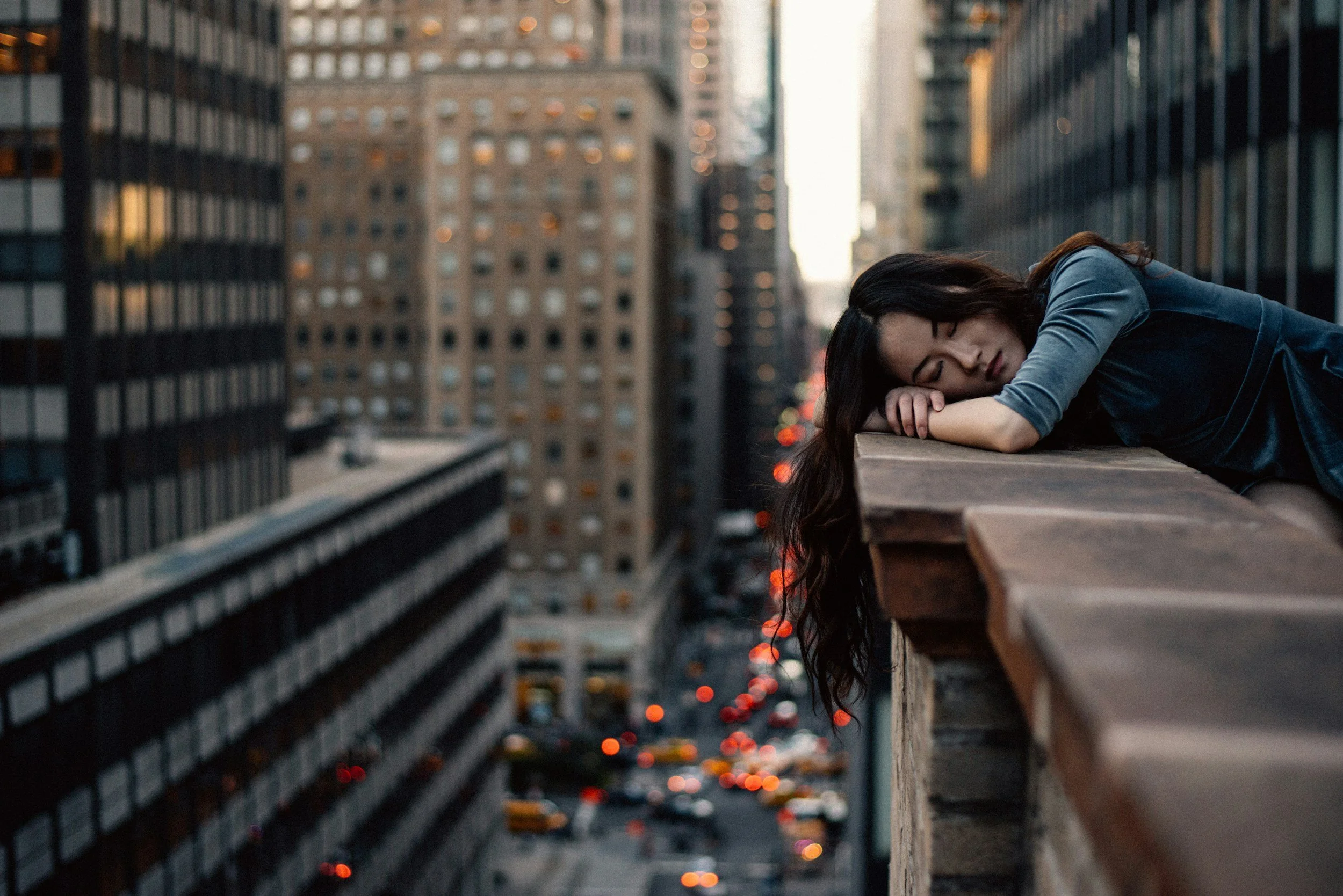 A woman with long dark hair lying on a rooftop ledge in a city at sunset, appearing to be asleep or exhausted.
