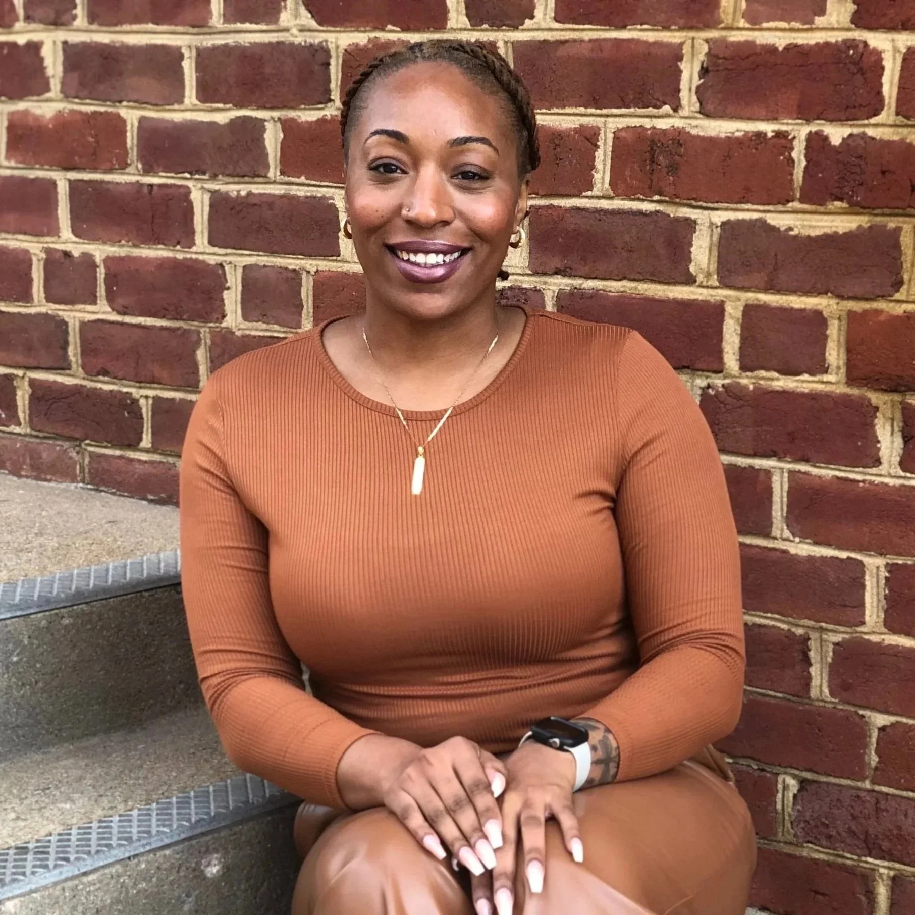 A woman with braided hair sitting on stairs in front of a brick wall, smiling, wearing a brown long-sleeve top, brown pants, a gold necklace, and a watch.
