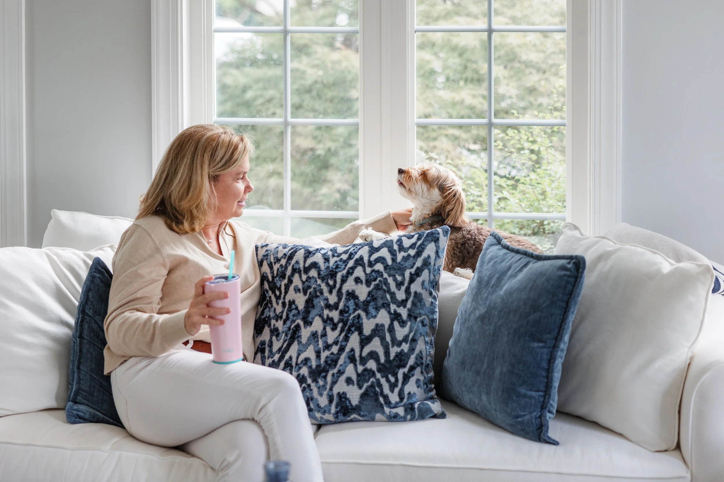A woman sitting on a white couch, holding a pink tumbler with a straw, and smiling at a small dog sitting on a pillow near her. The setting is a bright living room with large windows and greenery outside.