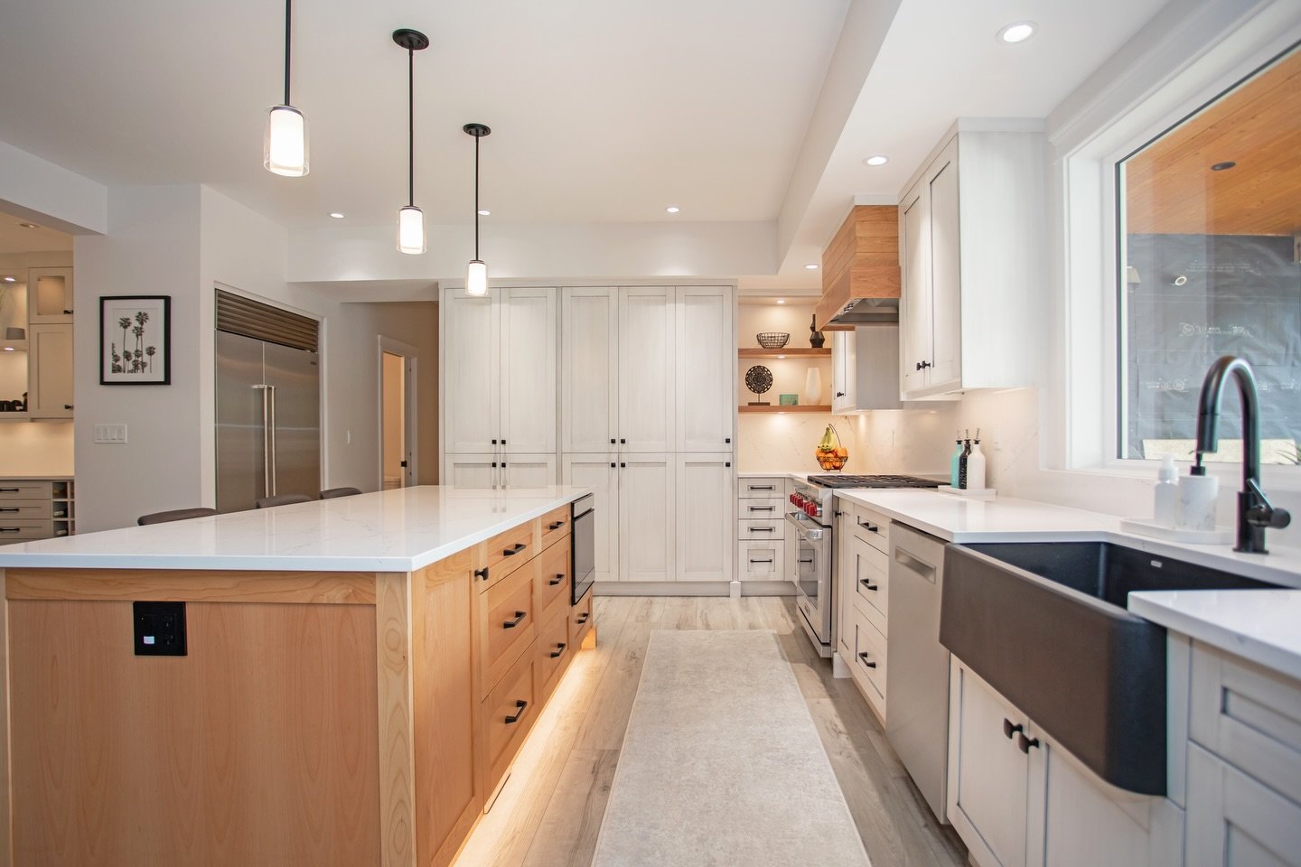 Two-toned kitchens remain one of the most effective ways to introduce contrast while preserving a sense of cohesion.
In this renovation, the hood surround, island, and open corner shelving are rendered in a light wood finish that gently offsets the a