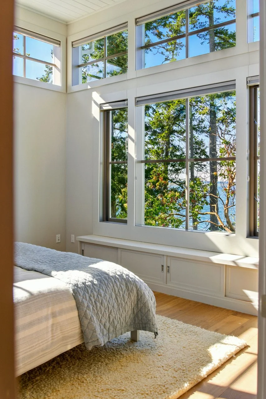 A bedroom with large windows overlooking trees and a view of the water, partly covered by white blinds, with sunlight casting shadows on the wall and a beige bed with a quilted blanket, cream-colored rug, and hardwood floor.
