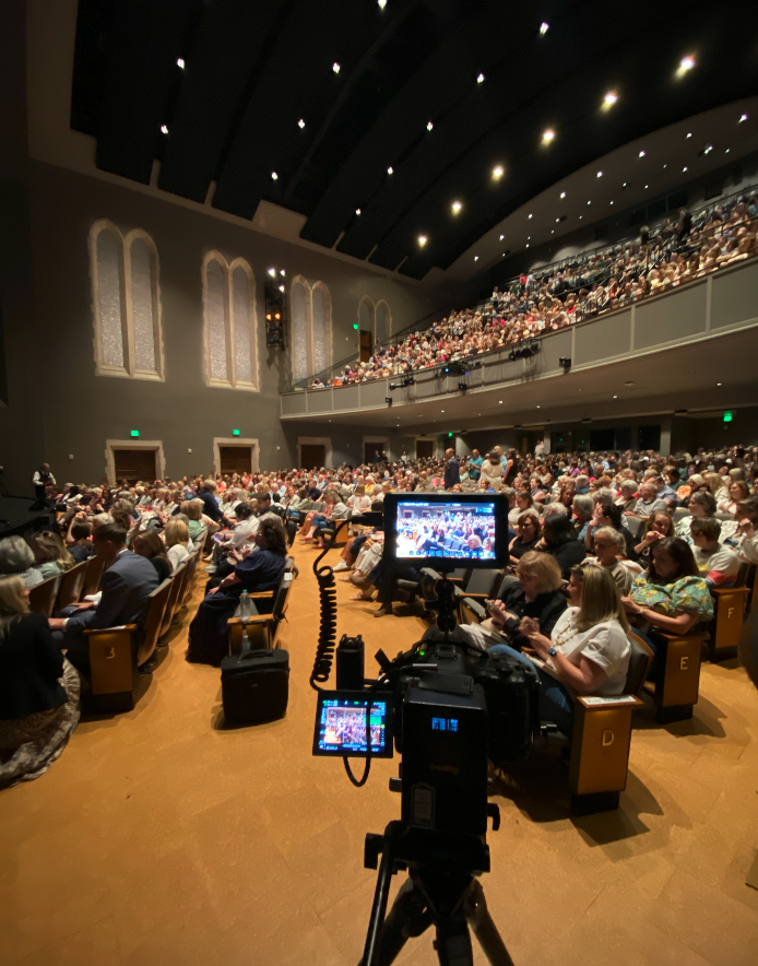 Audience filling a large auditorium while a professional video camera records from the aisle