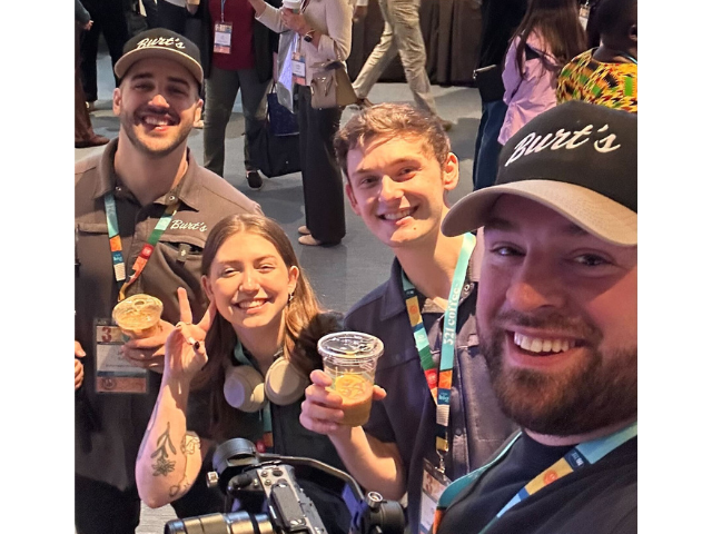 Four smiling people wearing event badges pose for a selfie at a crowded convention, holding drinks and wearing Burt’s hats.