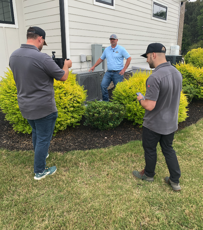 Three HVAC technicians filming an air conditioning unit outside a house