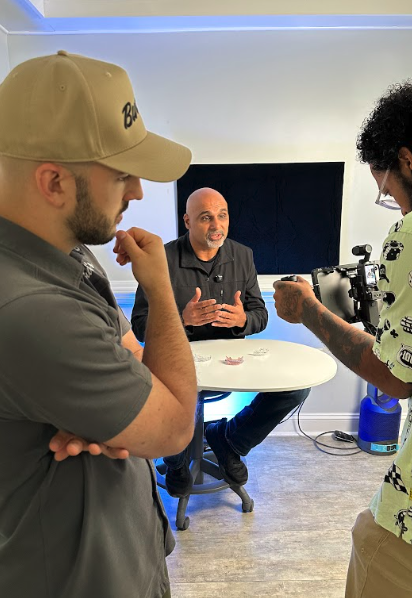 Behind-the-scenes video interview with a bald man speaking at a table while a camera operator films him in a studio
