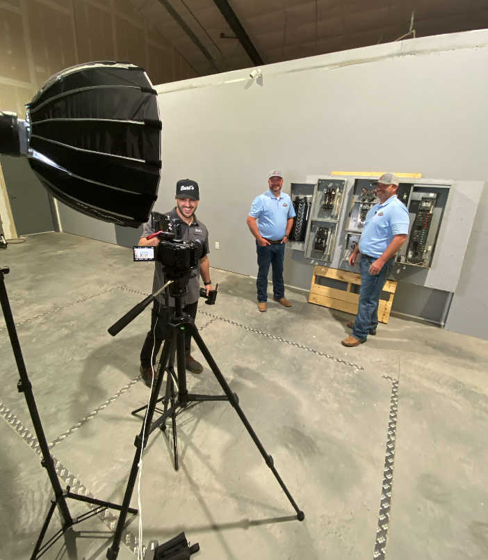 Two technicians pose beside industrial electrical panels while a cameraman films them in a warehouse.