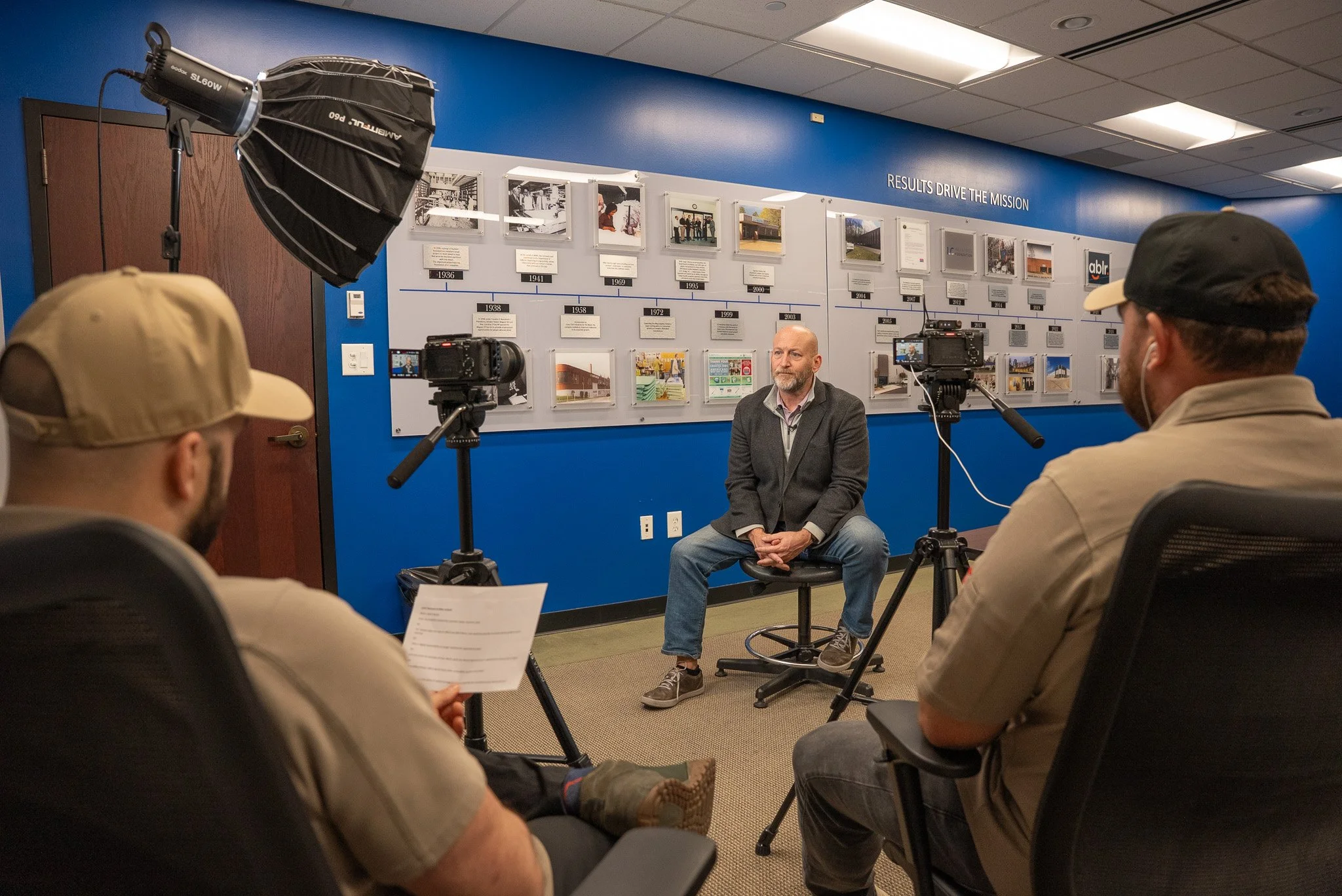 Film crew interviews a seated man in front of a corporate history wall using cameras and studio lighting.
