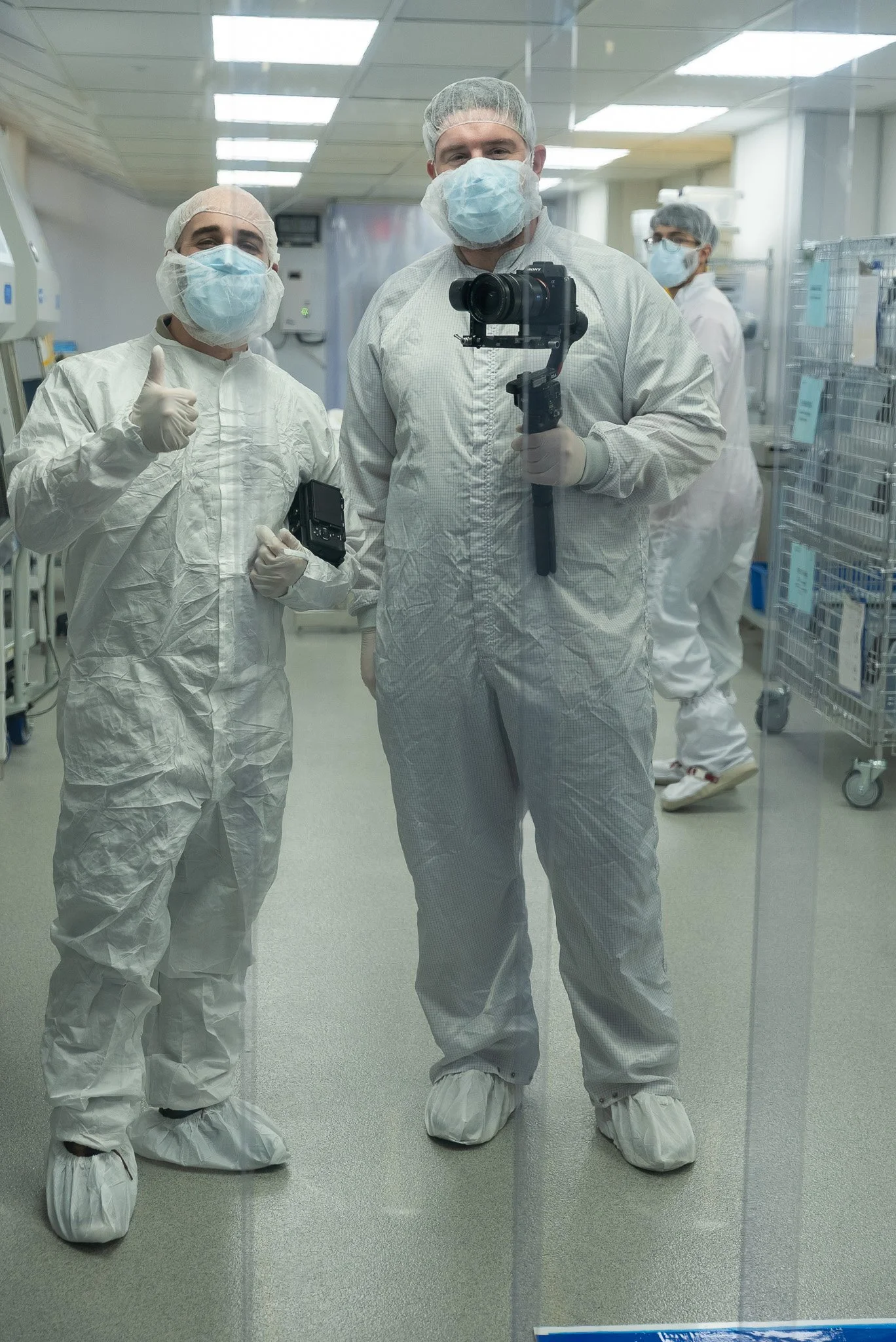 Two workers in full protective suits and masks stand in a cleanroom, with one holding a camera on a gimbal and the other giving a thumbs-up.