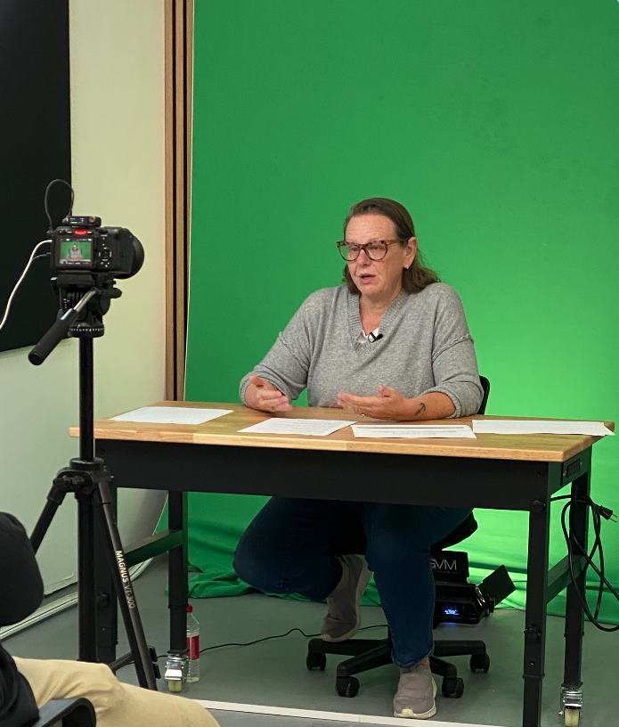 Woman speaking at a desk in front of a green screen while being recorded by a camera on a tripod