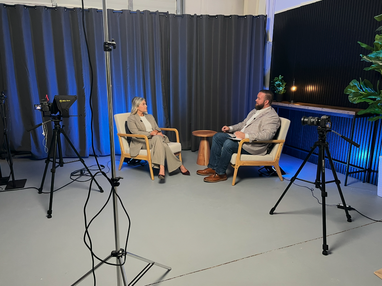Interview studio with two people seated in armchairs, cameras, lighting equipment, and gray curtains