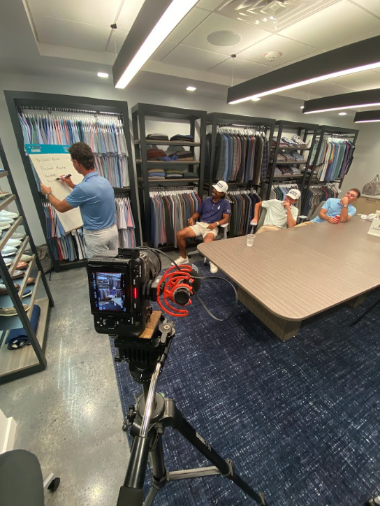 Video camera on a tripod filming a clothing store meeting with shirt racks, seated people, and a presenter at a whiteboard