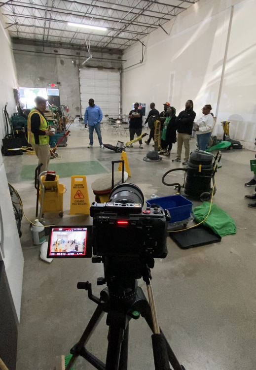 Video camera on a tripod filming a group of workers in a commercial cleaning equipment warehouse.