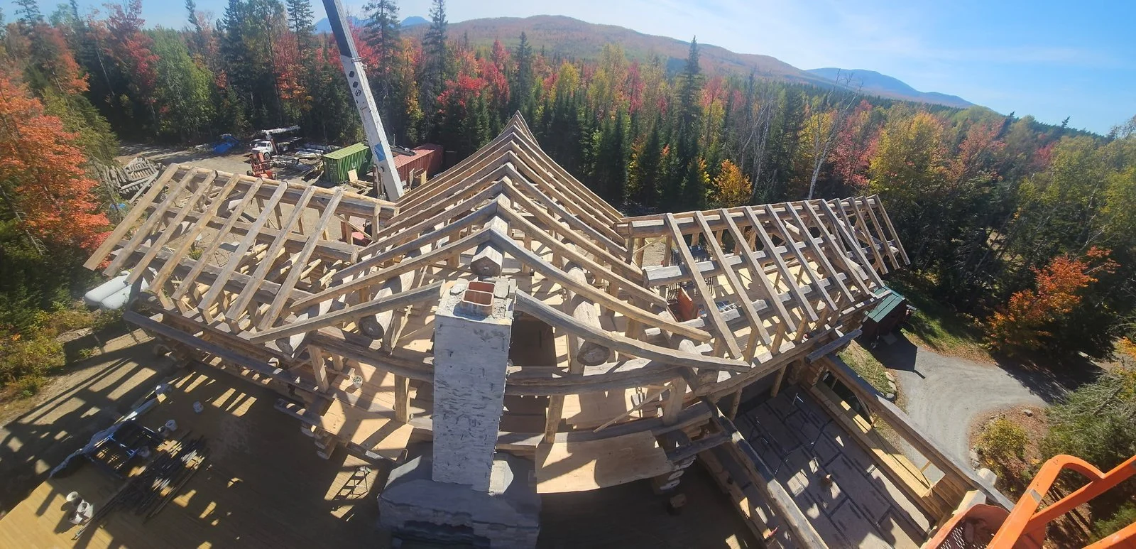 Elevated view of log home roof framing around a masonry chimney, surrounded by autumn trees.