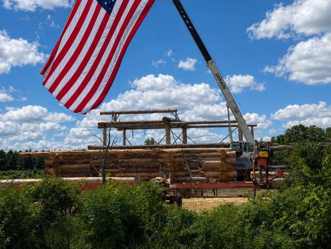 Crane and American flag above a log home shell with stacked walls and exposed beams.
