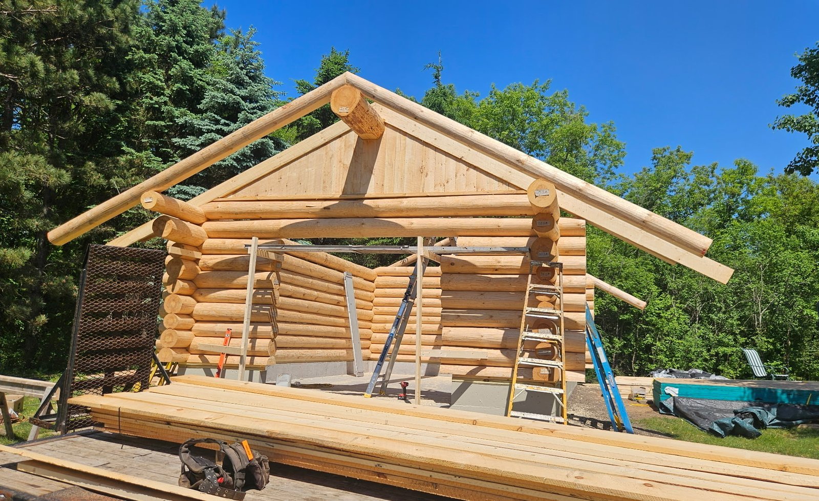 Front gable of a log shell during assembly, with fitted logs, roof beams, and ladders.
