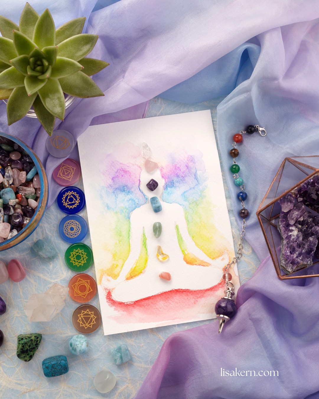 Watercolor painting of a meditating figure with chakra energy points displayed alongside healing crystals, tumbled stones, and a pendulum on a purple fabric background.