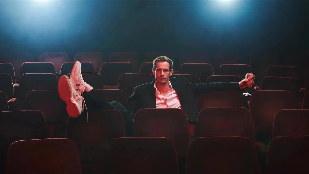 A man sitting alone in an empty theater or auditorium, with his legs crossed and relaxed, wearing a suit and holding a drink.