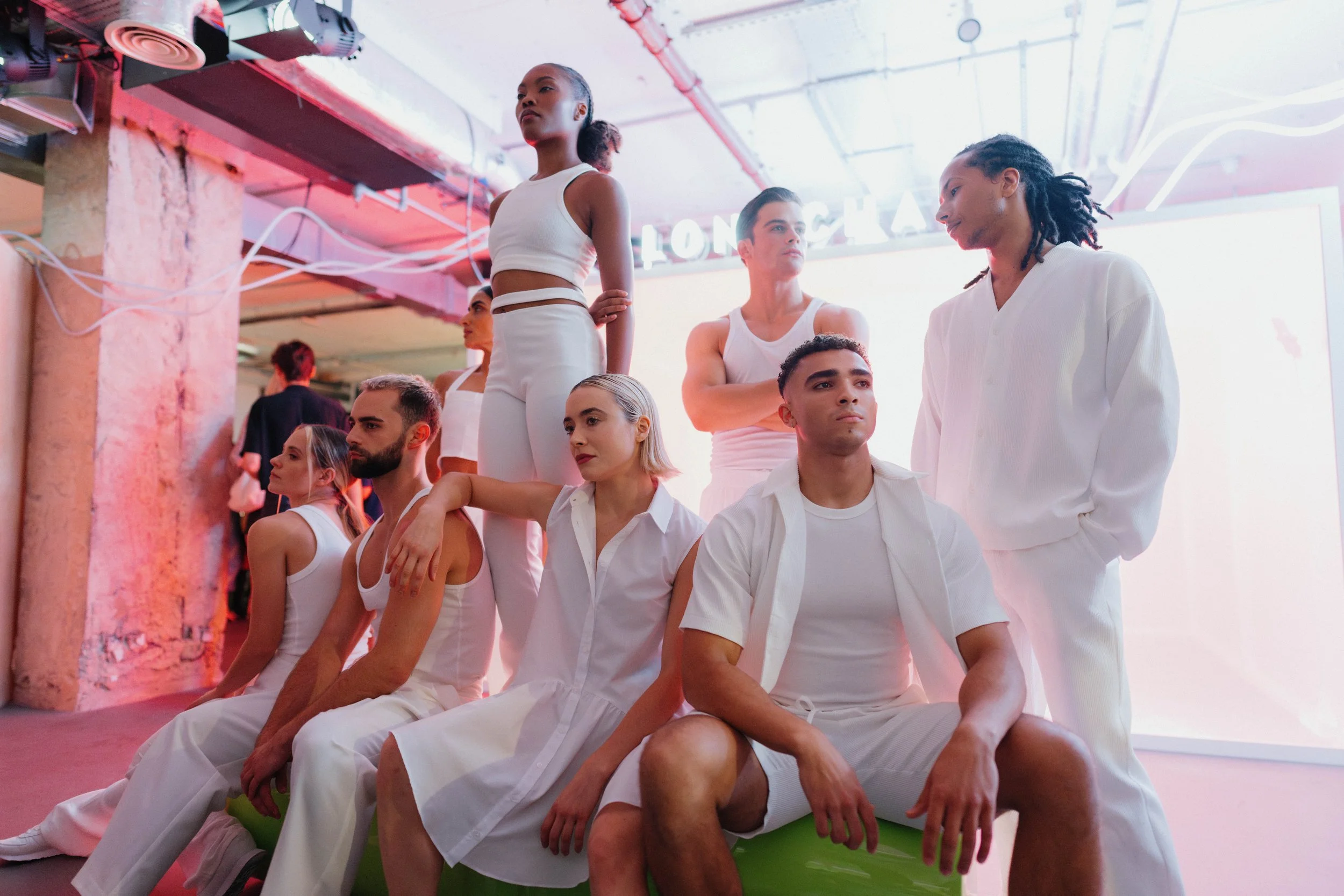 Group of diverse young adults dressed in white clothing, posing in a modern, industrial-style studio with pink neon lights.