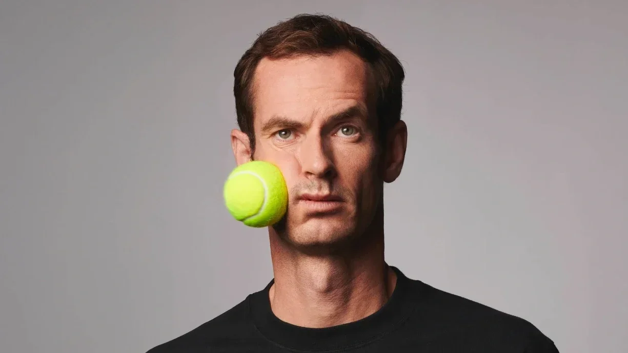 Andy Murray with a tennis ball on his face, positioned near his cheek.