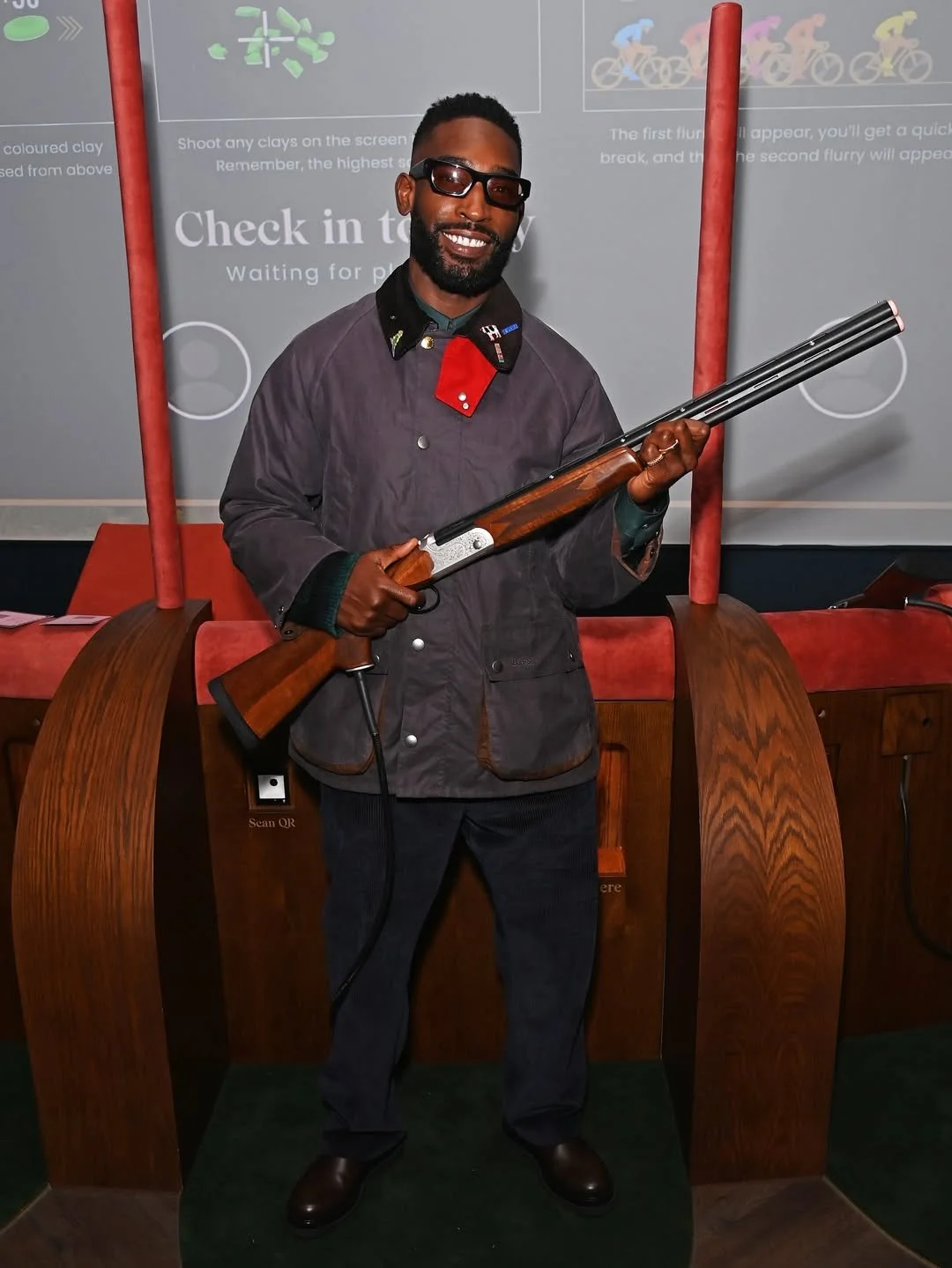A man smiling and wearing glasses, holding a double-barreled shotgun, standing in front of a background with cycling icons and instructions.