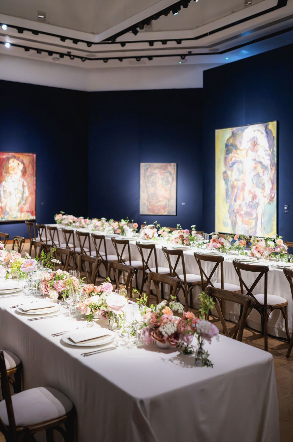 A decorated banquet table with pink and white flowers, set for a formal event in an art gallery with dark blue walls and colorful paintings.