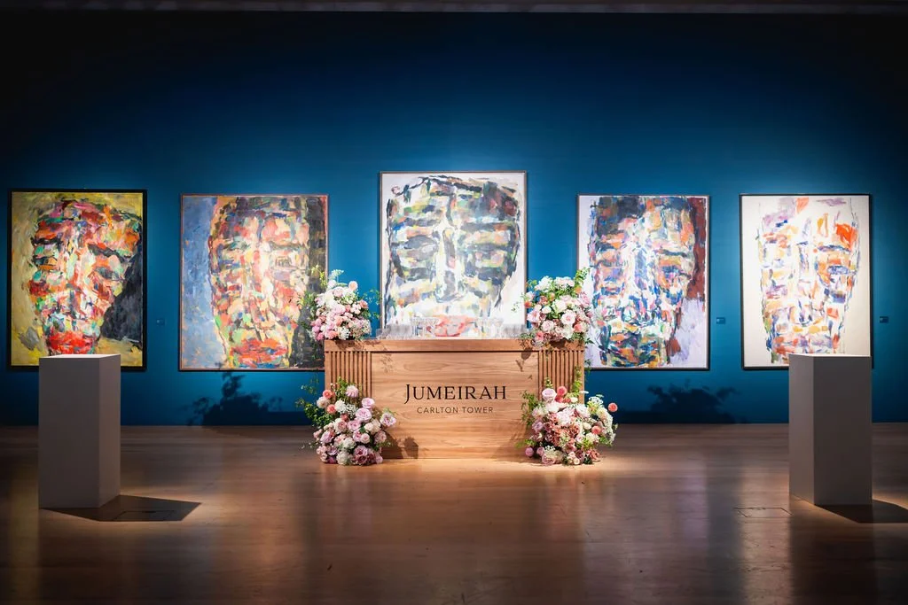 An art gallery with five abstract portrait paintings on a blue wall, a wooden pedestal with the word 'Jumeirah' and floral arrangements in front of it.