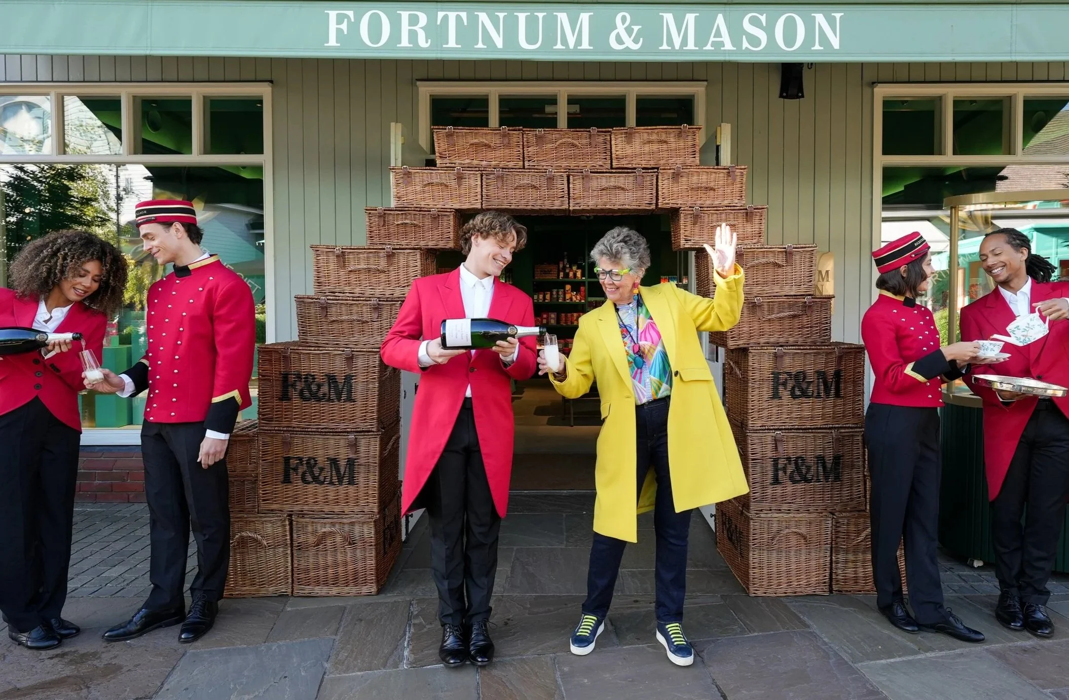 People dressed as hotel bellhops celebrating outside P&M store with wicker baskets and a sign reading "Fortnum & Mason."