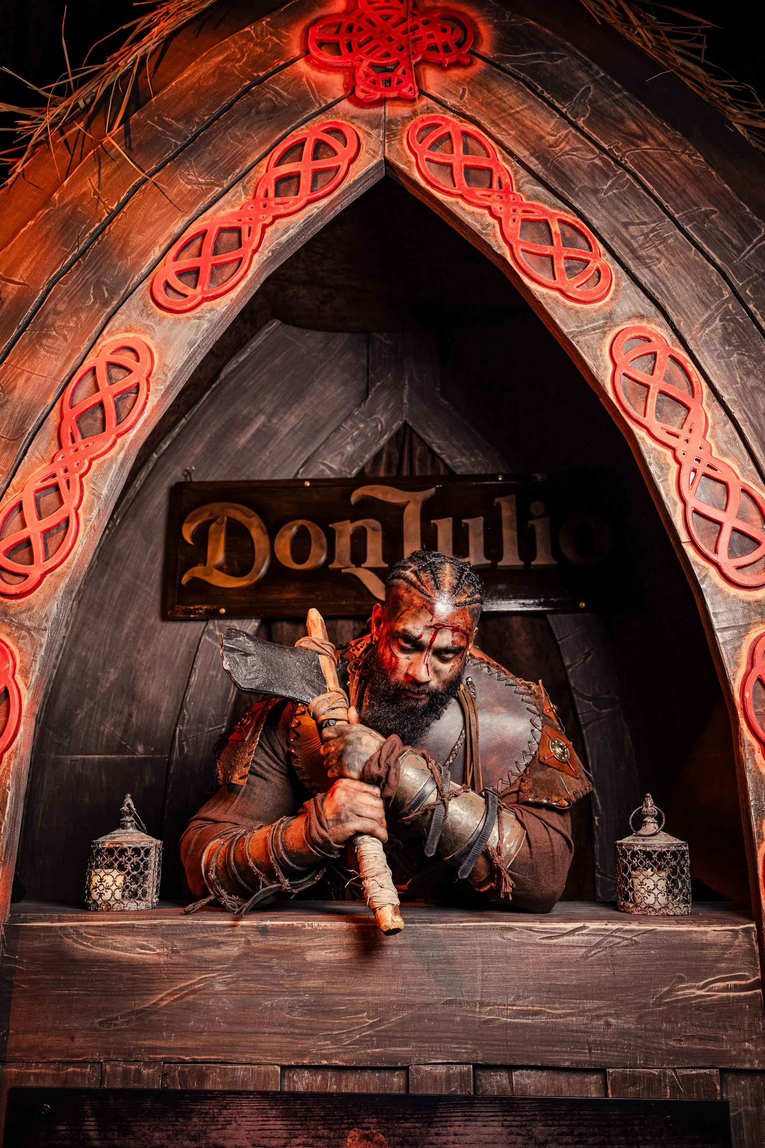 A person dressed as a medieval or fantasy warrior with face paint, holding a large axe, sitting behind a wooden counter under a gothic-style arch with red decorative accents, and a sign that reads 'Don Julio' in the background.