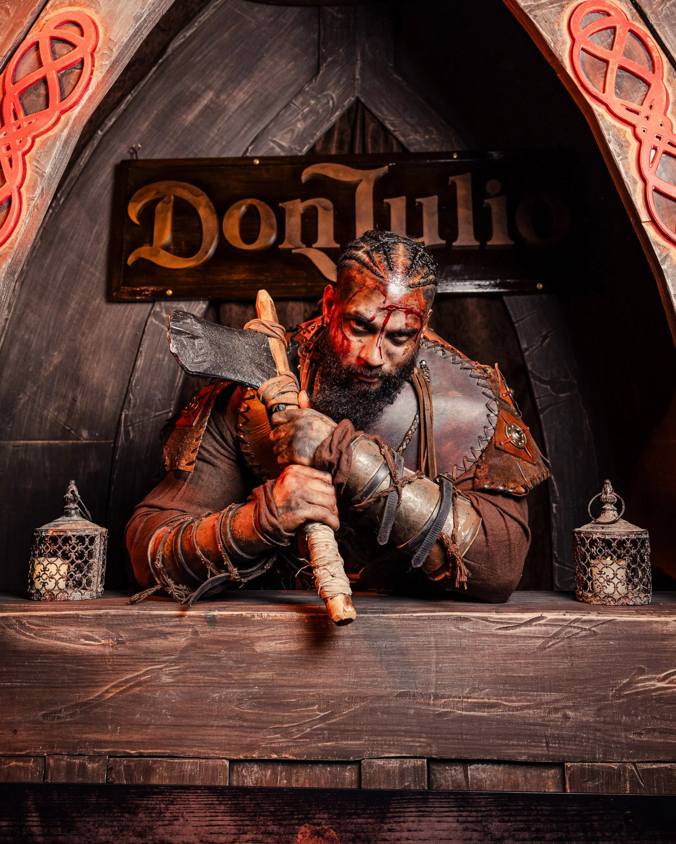 A man with a beard and braided hair, dressed in medieval-style leather armor, sitting behind a wooden counter with two lit lanterns, holding a large axe with both hands, in front of a dark wooden backdrop with a sign that says 'Don Julio'.