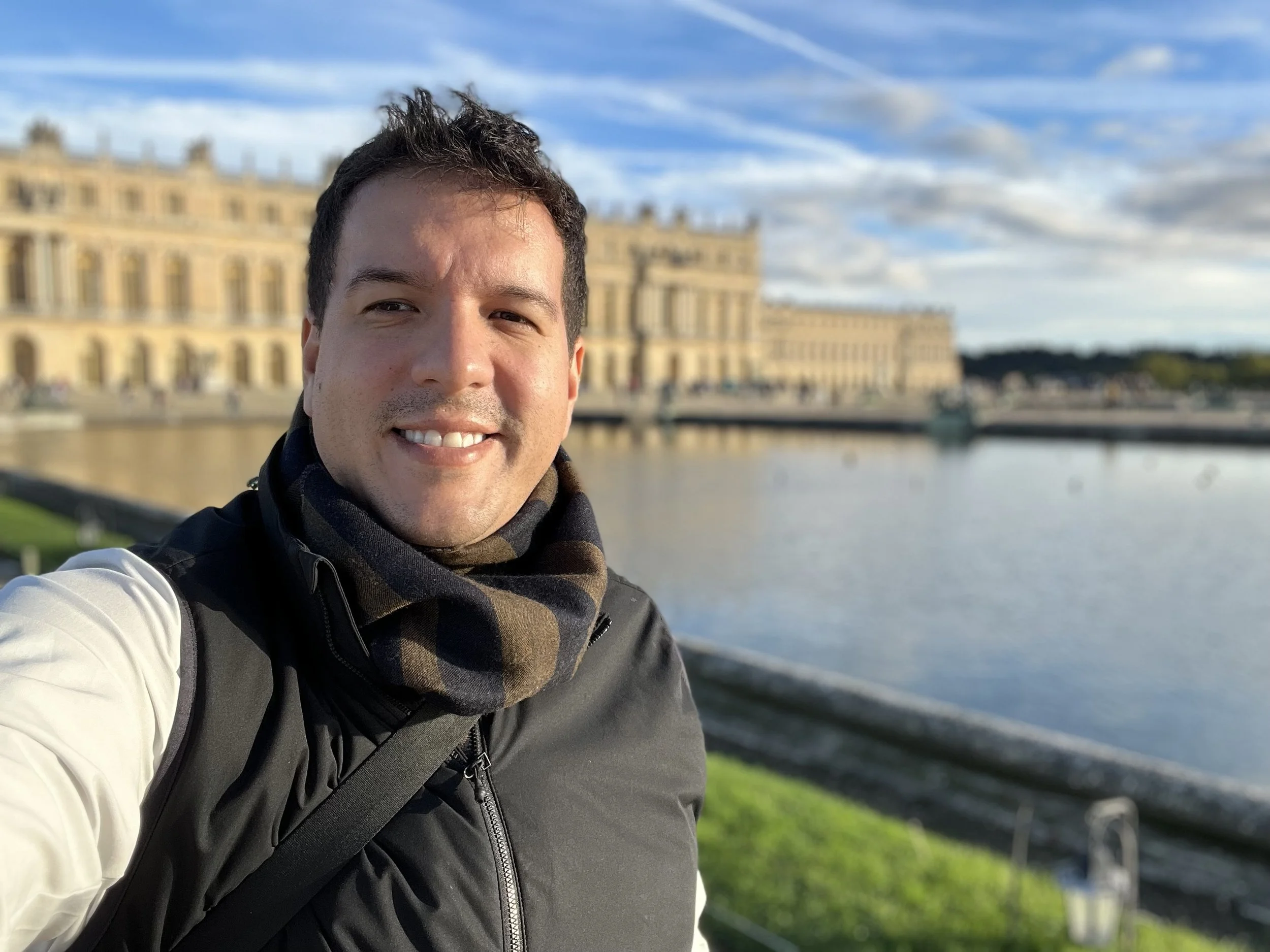 Joven sonriente tomando una selfie en un parque junto a un río, con un edificio histórico en el fondo y un cielo con nubes.