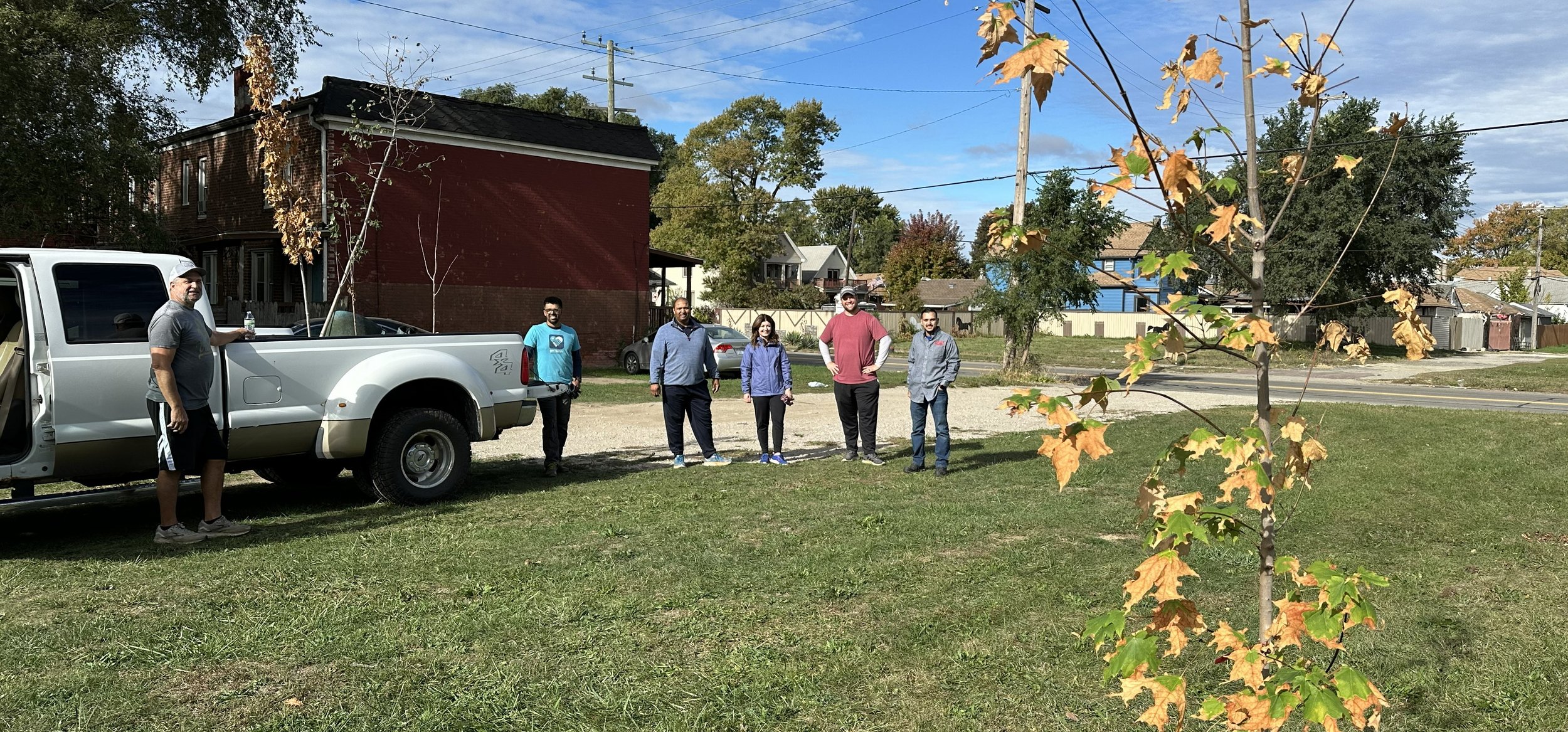Group of six people standing on grass in a park or yard, with a white pickup truck parked nearby, a red brick house in the background, and trees with fall foliage.