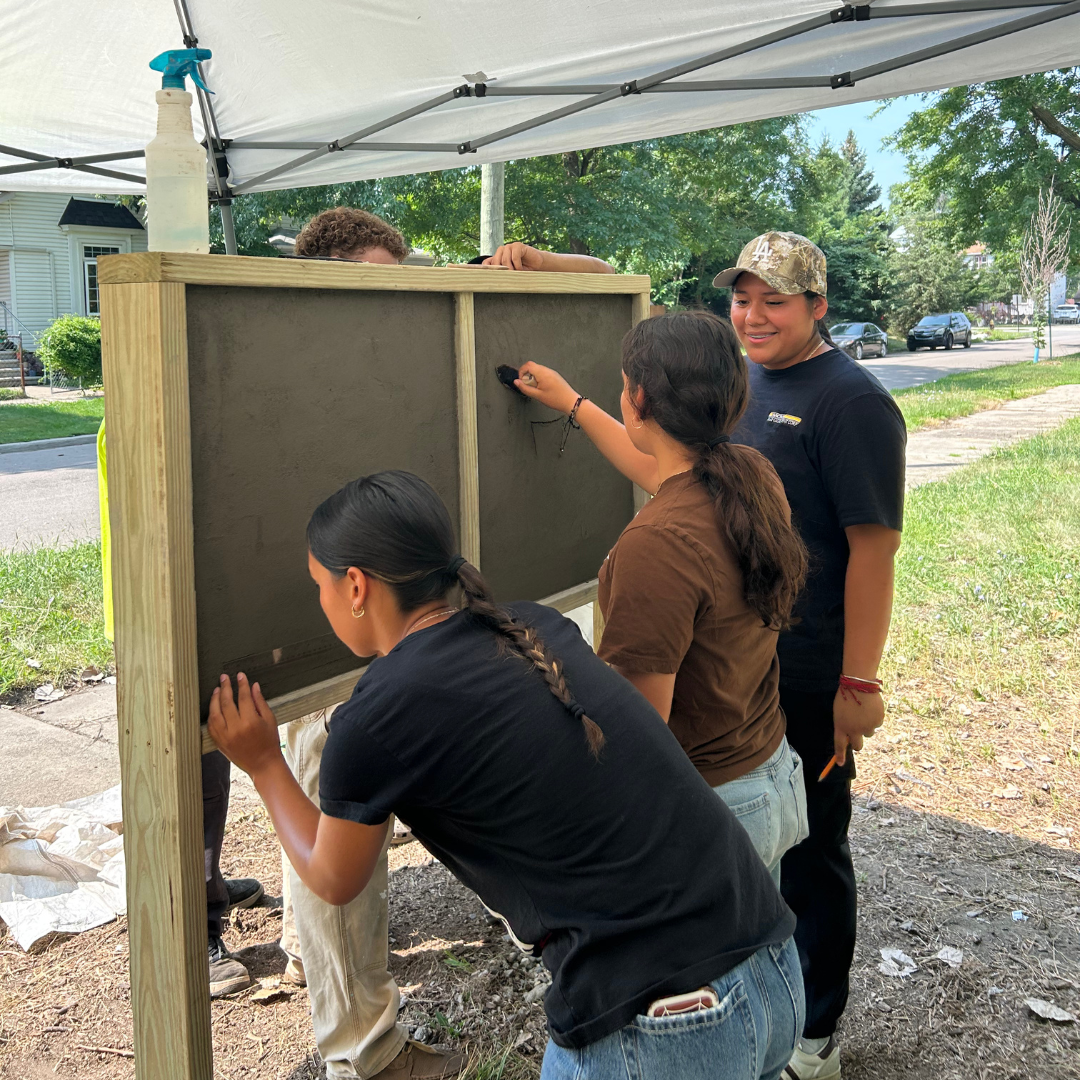 Four people working together to paint a large wooden frame with gray concrete. They are outdoors under a canopy, with trees and a street visible in the background.