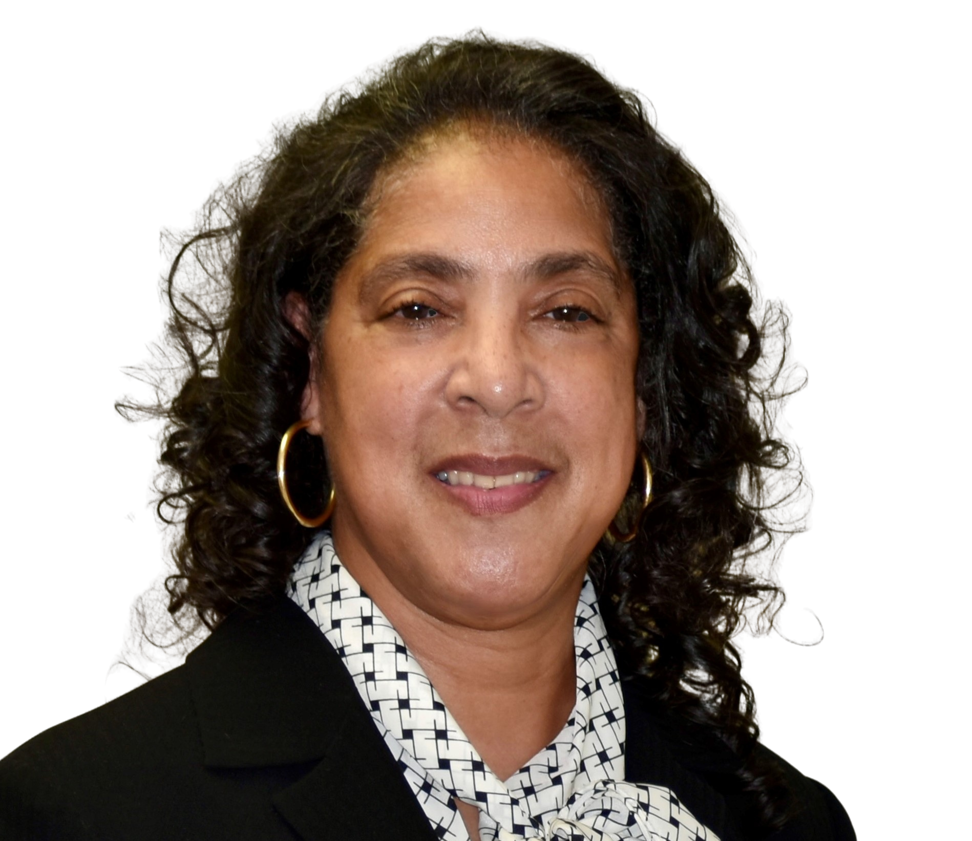 Portrait of a woman with dark curly hair, wearing gold hoop earrings, a black blazer, and a patterned white and black blouse, smiling at the camera.