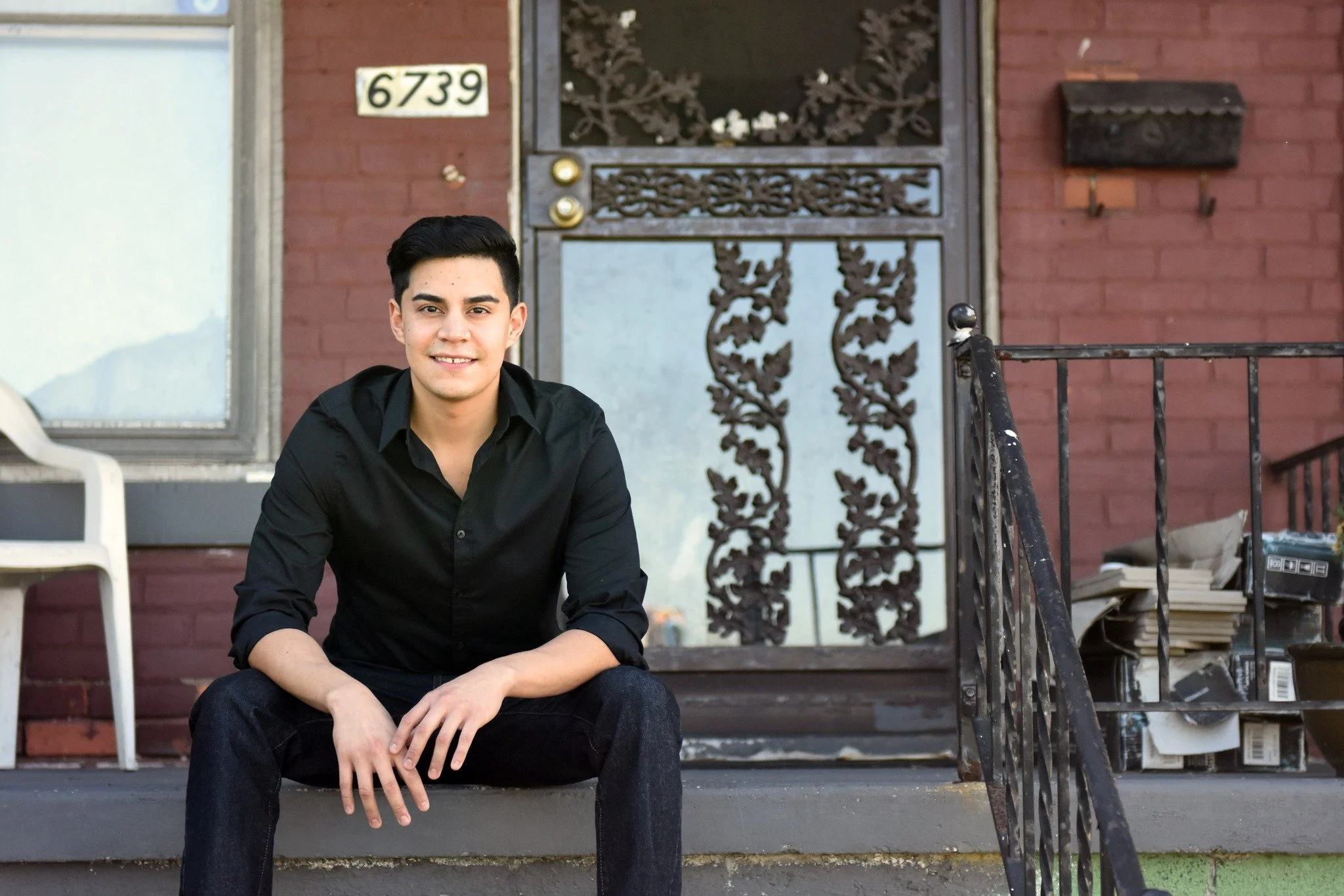 A young man with black hair, wearing a black shirt and jeans, sitting on the front steps of a house with a brick exterior, a decorative metal door, and a white plastic chair nearby.