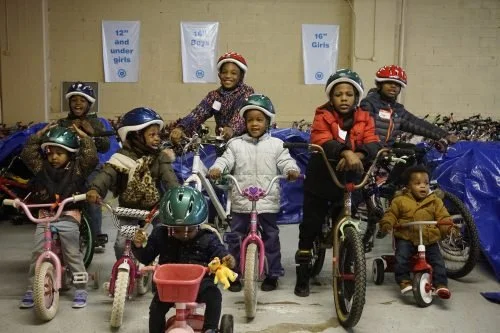 Group of young children wearing bike helmets, riding bicycles and tricycles indoors, with some standing and others sitting on their bikes, against a backdrop of blue bags and signs.