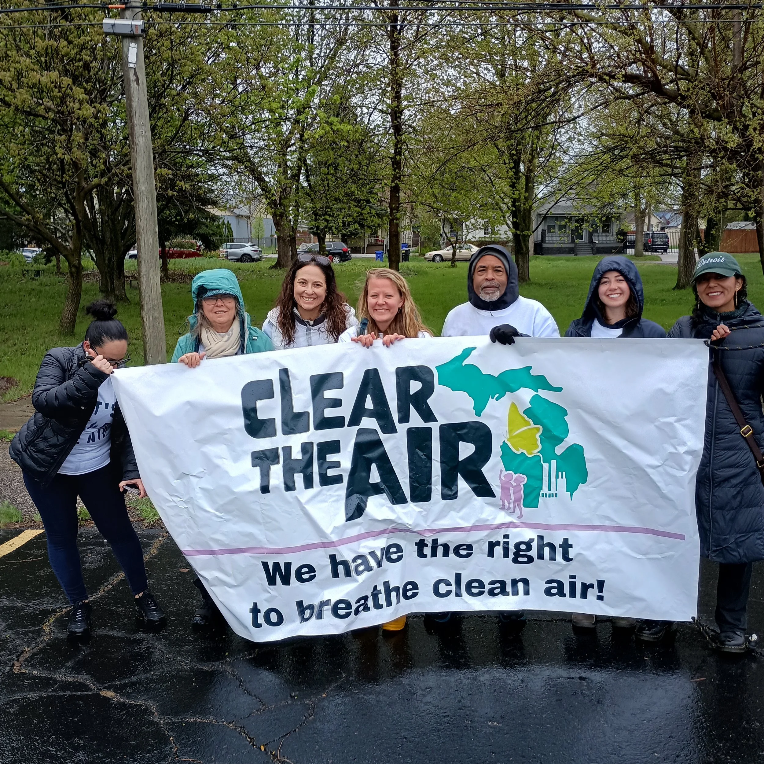 Group of people holding a banner that reads "Clear the Air - We have the right to breathe clean air!" during an outdoor event with trees and houses in the background.