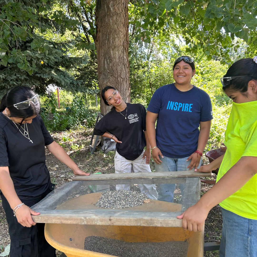 Four women working outdoors on a project involving sand and small stones, standing around a wooden frame and mesh, smiling and wearing safety glasses, with trees and greenery in the background.