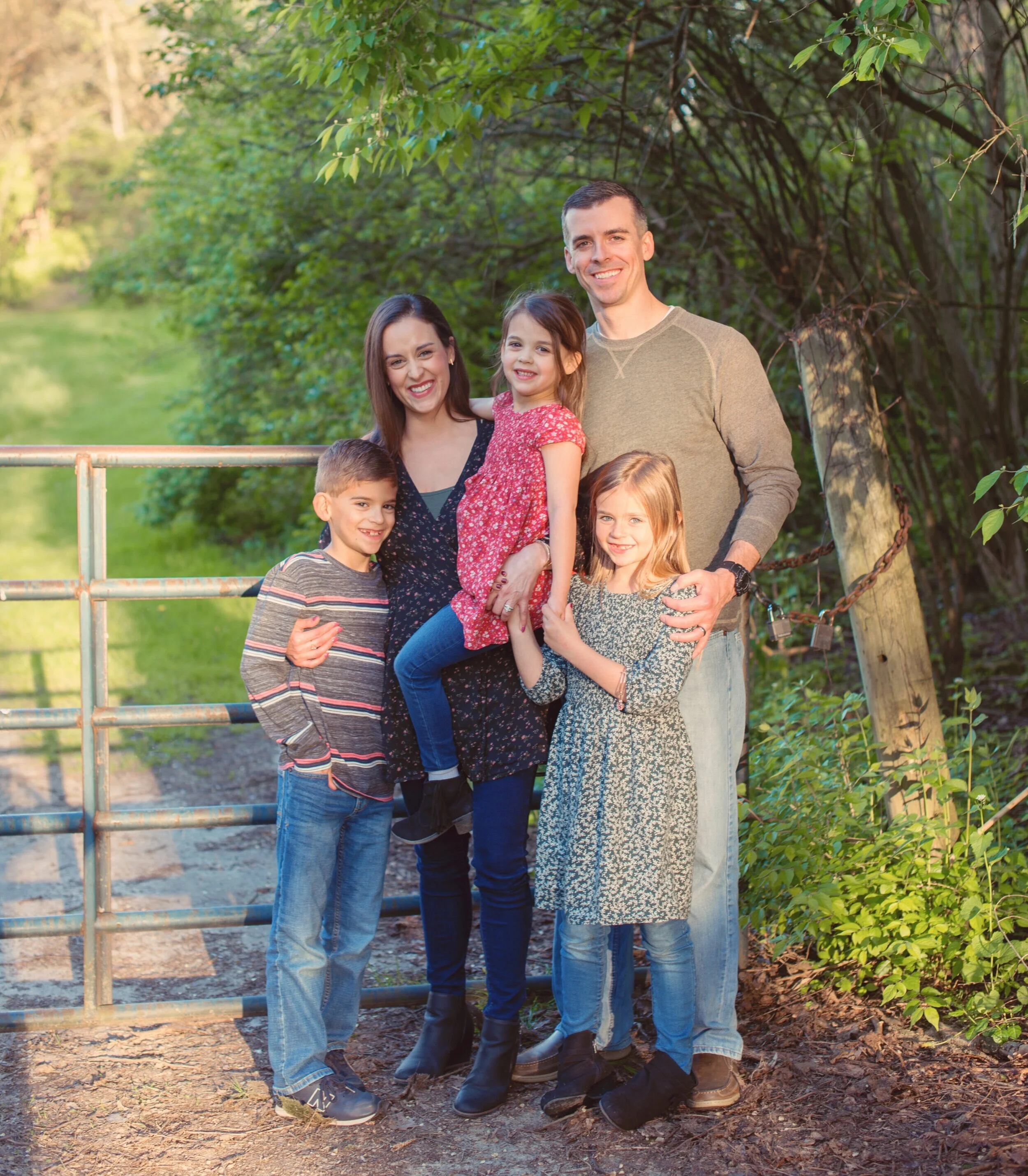 A family of six standing outdoors near a metal gate surrounded by trees and greenery, smiling at the camera.